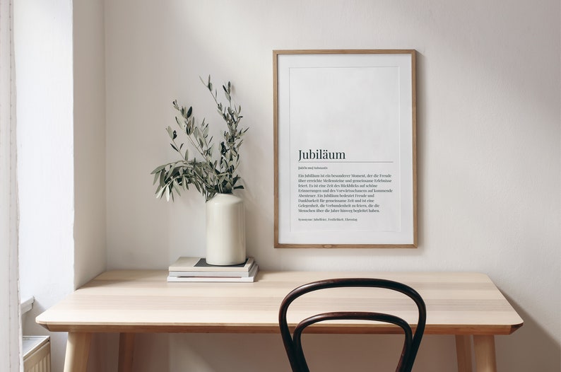 May include: A light wood desk with a framed print that reads "Jubil&auml;um" in black text. The print is on a white background. A vase with green branches is on the desk, along with a stack of books.