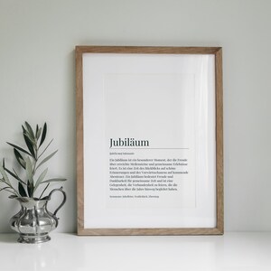 May include: A framed print with a white background and black text. The text reads "Jubil&auml;um" and a definition of the word in German. The frame is made of light brown wood.