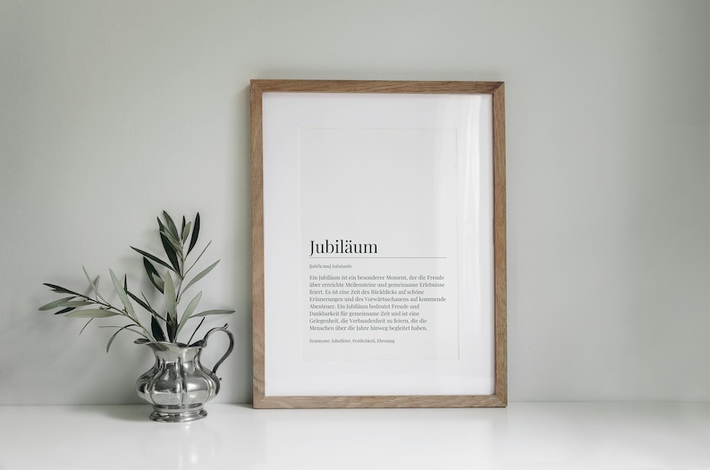 May include: A framed print with a white background and black text. The text reads "Jubil&auml;um" and a definition of the word in German. The frame is made of light brown wood.