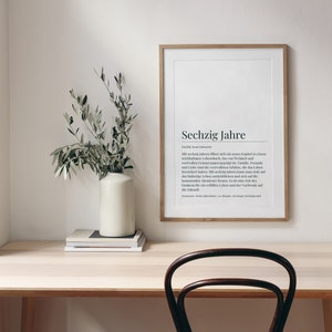 May include: A framed print with the German word "Sechzig Jahre" and a definition in German. The print is hanging on a white wall above a light wood desk with a vase of green branches and a stack of books. A dark brown chair is in front of the desk.