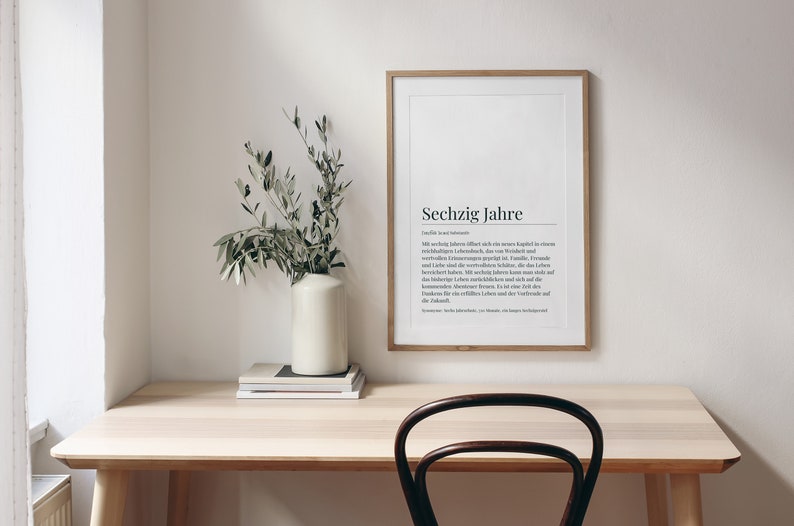 May include: A framed print with the German word "Sechzig Jahre" and a definition in German. The print is hanging on a white wall above a light wood desk with a vase of green branches and a stack of books. A dark brown chair is in front of the desk.