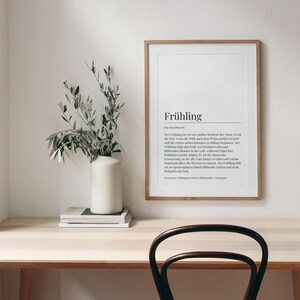 May include: A framed print with the German word "Fr&uuml;hling" (Spring) and its definition in German. The print is hanging on a white wall above a wooden desk with a vase of green branches and a stack of books.