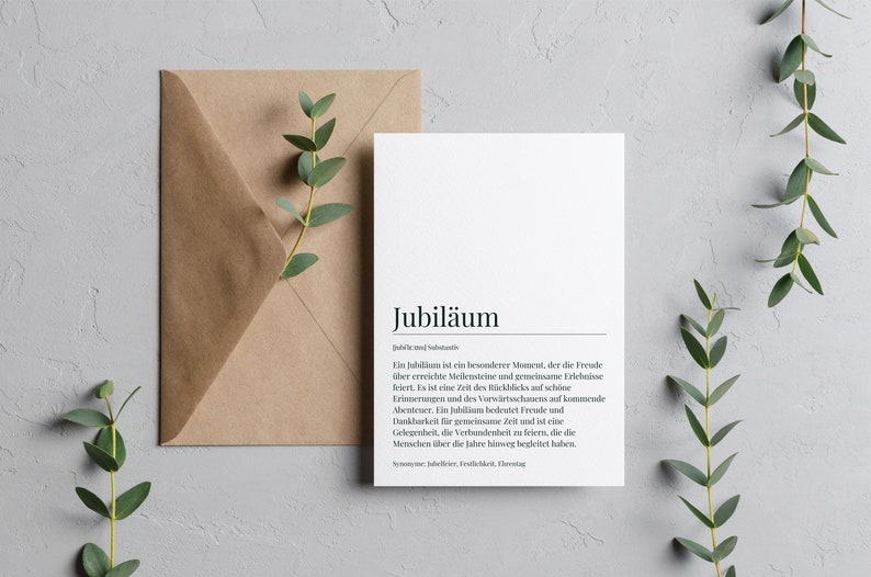 May include: A white card with the word "Jubil&auml;um" in black text. The card defines the word "Jubil&auml;um" in German, explaining that it is a special moment that celebrates milestones and shared experiences. The card is on a gray background with green foliage.