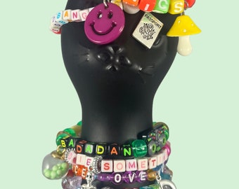 May include: A set of colourful beaded bracelets with letter charms spelling out phrases like "SUBTRONICS", "BAD AND DICE", "SOMETIME", and "LOVE HARD". The bracelets also feature a smiley face charm, a passport charm, a mushroom charm, a skeleton charm, and a music note charm.