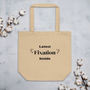 May include: A beige tote bag with black text that reads "Latest Fixation Inside" and two small diamond shapes on either side of the word "Fixation".