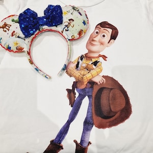 Op de afbeelding: Een wit T-shirt met een kleurrijk cartoonbeeld van Woody uit Toy Story. Woody draagt zijn kenmerkende cowboyhoed en vest. Hij houdt een blauw en wit hoofdband met een strik vast. Het hoofdband heeft een cartoonontwerp met verschillende personages uit Toy Story.