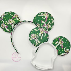 May include: A headband featuring two padded circular ears covered in green fabric with a repeating pattern of Buzz Lightyear characters and yellow stars. The headband has a white elastic strap attached to the bottom for secure placement.