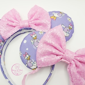 May include: A headband featuring two round ears covered in a light purple fabric with a repeating Daisy Duck and white flower pattern. Each ear is adorned with a large, light pink sequined bow. A circular logo reads Bow U Tiful Bow-Tique.