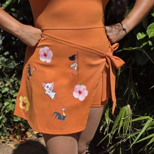 Op de afbeelding: Een oranje overslagrok met ingebouwde short, voorzien van een striksluiting aan de zijkant en een print van een cartoonmeisje, een varken, een haan en roze en gele hibiscusbloemen. Het kledingstuk is gemaakt van een gladde, effen stof.