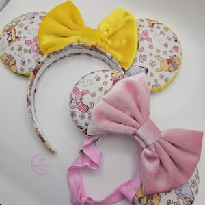 May include: Two headbands featuring circular ears with a white fabric pattern of cartoon characters including a bear, a pig, and a donkey. One headband has a bright yellow velvet bow, while the other has a soft pink velvet bow and a pink elastic strap.