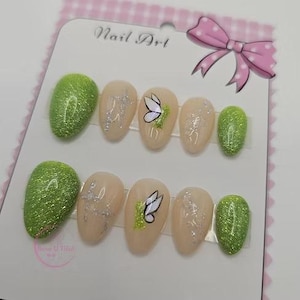 Può includere: Un set di unghie finte ovali. Le unghie presentano un mix di verde glitterato e nude, con dettagli argentati e disegni di farfalle. Il testo "Nail Art" è visibile su sfondo rosa chiaro. Presentato su una carta.
