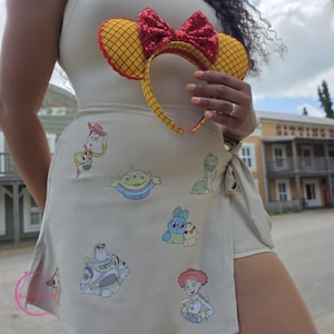 May include: A beige wrap skirt adorned with colorful Toy Story character decals. A matching headband with yellow ears and a red sequined bow is held up. The skirt features a side slit and a tie closure.