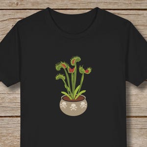 May include: Black t-shirt with a graphic of a potted Venus flytrap plant. The pot is decorated with white skull and crossbones.