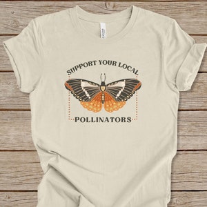 Puede incluir: Una camiseta beige con un gráfico de una mariposa y el texto "Apoya a tus polinizadores locales".