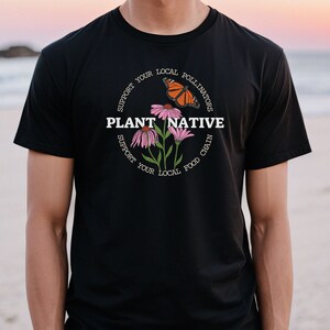Pode incluir: Camiseta preta com um gr&aacute;fico de flores cor-de-rosa e uma borboleta. O gr&aacute;fico &eacute; rodeado por texto que diz "Apoie os seus polinizadores locais Plante nativas Apoie a sua cadeia alimentar local".
