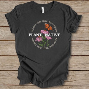 Pode incluir: Uma camiseta cinza escura com um gr&aacute;fico circular branco que apresenta o texto "Plantar Nativo" e as palavras "Apoie seus polinizadores locais" e "Apoie sua cadeia alimentar local" ao redor do c&iacute;rculo. O gr&aacute;fico tamb&eacute;m inclui uma borboleta e flores cor-de-rosa.