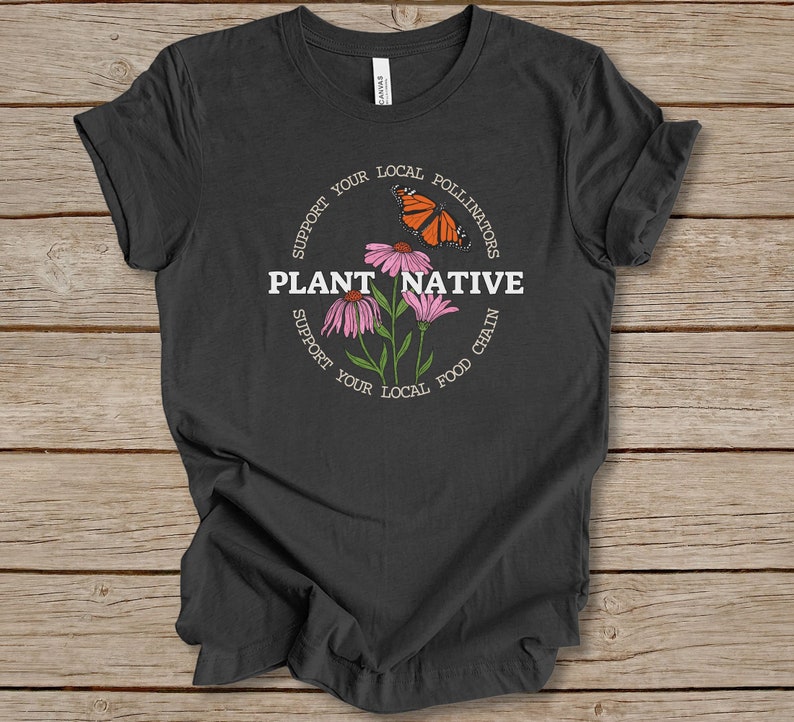 Pode incluir: Uma camiseta cinza escura com um gr&aacute;fico circular branco que apresenta o texto "Plantar Nativo" e as palavras "Apoie seus polinizadores locais" e "Apoie sua cadeia alimentar local" ao redor do c&iacute;rculo. O gr&aacute;fico tamb&eacute;m inclui uma borboleta e flores cor-de-rosa.