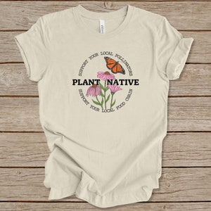 Pode incluir: Uma camiseta bege com um design circular que apresenta uma borboleta monarca, flores cor-de-rosa e o texto "Apoie seus polinizadores locais Plante nativas Apoie sua cadeia alimentar local".
