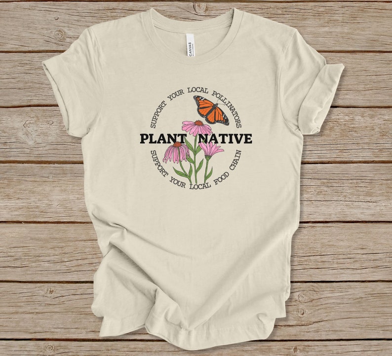 Pode incluir: Uma camiseta bege com um design circular que apresenta uma borboleta monarca, flores cor-de-rosa e o texto "Apoie seus polinizadores locais Plante nativas Apoie sua cadeia alimentar local".