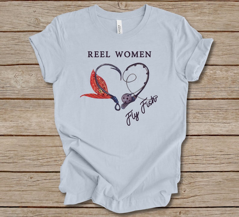 May include: Light blue t-shirt with the text "Reel Women Fly Fish" in black lettering. The text is surrounded by a heart shape made of a fishing hook and a fly fishing lure.
