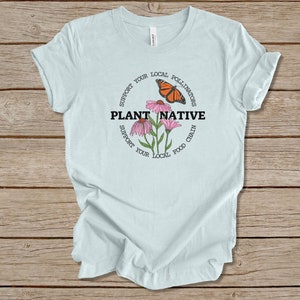 Pode incluir: Camiseta azul claro com um design circular que apresenta uma borboleta monarca, flores cor-de-rosa e o texto "PLANT NATIVE" em preto. O c&iacute;rculo tamb&eacute;m inclui o texto "SUPPORT YOUR LOCAL POLLINATORS" e "SUPPORT YOUR LOCAL FOOD CHAIN".