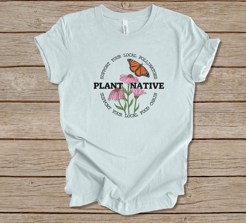 Pode incluir: Camiseta azul claro com um design circular que apresenta uma borboleta monarca, flores cor-de-rosa e o texto "PLANT NATIVE" em preto. O c&iacute;rculo tamb&eacute;m inclui o texto "SUPPORT YOUR LOCAL POLLINATORS" e "SUPPORT YOUR LOCAL FOOD CHAIN".