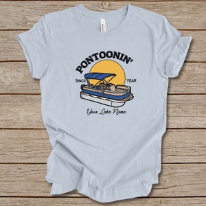 K&ouml;nnte beinhalten: Hellblaues T-Shirt mit einem Grafikdruck eines Pontonboots auf einem See mit einer gelben Sonne und dem Text "Pontoonin' Since Year" und "Your Lake Name".
