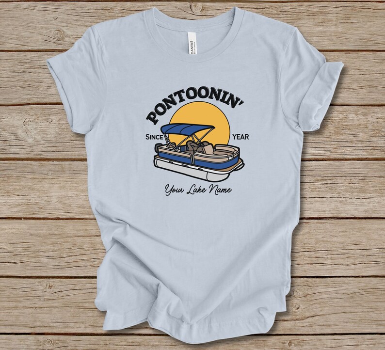 K&ouml;nnte beinhalten: Hellblaues T-Shirt mit einem Grafikdruck eines Pontonboots auf einem See mit einer gelben Sonne und dem Text "Pontoonin' Since Year" und "Your Lake Name".