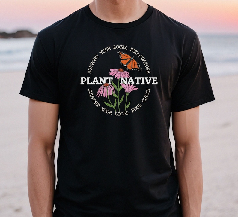 Pode incluir: Camiseta preta com um gr&aacute;fico de flores cor-de-rosa e uma borboleta. O gr&aacute;fico &eacute; rodeado por texto que diz "Apoie os seus polinizadores locais Plante nativas Apoie a sua cadeia alimentar local".