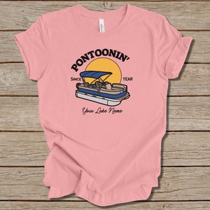 K&ouml;nnte beinhalten: Rosa T-Shirt mit einem Grafikdruck eines Pontonboots auf einem See mit einer Sonne und dem Text "Pontoonin' Since Year" und "Your Lake Name".
