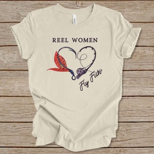 May include: A beige t-shirt with the text "Reel Women Fly Fish" in black. The text is inside a heart shape made of two fishing hooks and a red and orange fly.