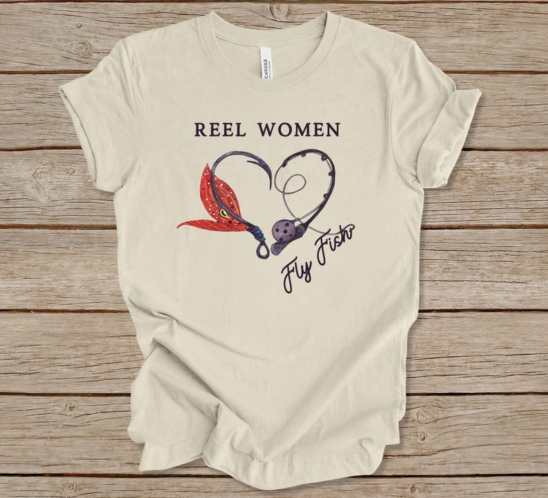May include: A beige t-shirt with the text "Reel Women Fly Fish" in black. The text is inside a heart shape made of two fishing hooks and a red and orange fly.
