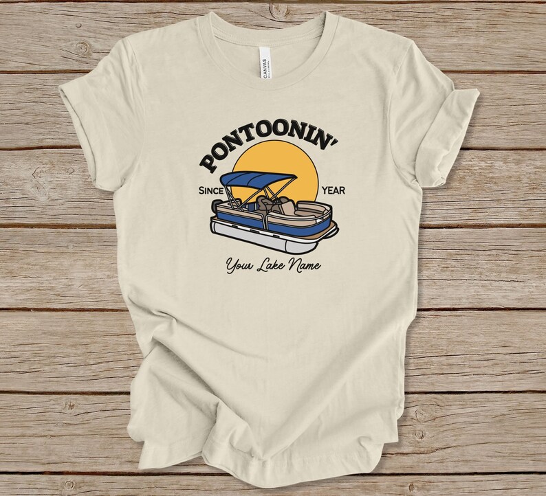 K&ouml;nnte beinhalten: Ein beiges T-Shirt mit einem Aufdruck eines Pontonboots mit Verdeck, einer gelben Sonne und dem Text "Pontoonin' Since YEAR". Der Text "Your Lake Name" befindet sich unter dem Aufdruck.