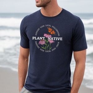Pode incluir: Uma camiseta azul marinho com um gr&aacute;fico circular branco que apresenta uma borboleta, flores e o texto "PLANT NATIVE" com o texto "SUPPORT YOUR LOCAL POLLINATORS" e "SUPPORT YOUR LOCAL FOOD CHAIN" que circunda o gr&aacute;fico.