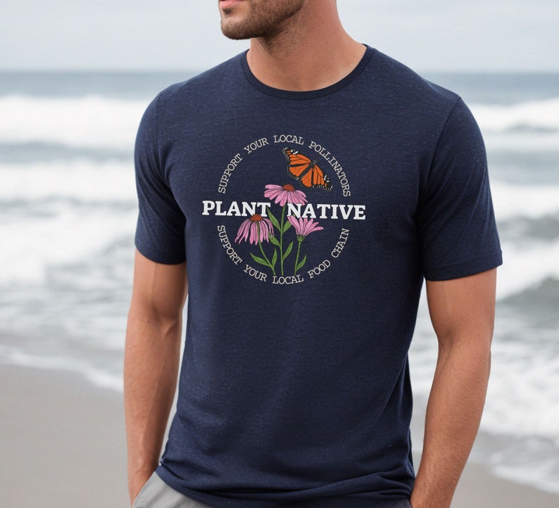 Pode incluir: Uma camiseta azul marinho com um gr&aacute;fico circular branco que apresenta uma borboleta, flores e o texto "PLANT NATIVE" com o texto "SUPPORT YOUR LOCAL POLLINATORS" e "SUPPORT YOUR LOCAL FOOD CHAIN" que circunda o gr&aacute;fico.