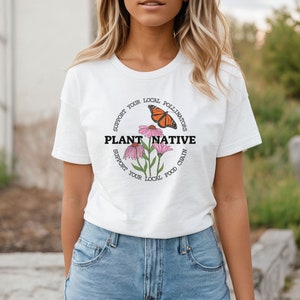 Pode incluir: Camiseta branca com um design gr&aacute;fico circular que apresenta uma borboleta, flores e o texto "Apoie seus polinizadores locais Plante nativos Apoie sua cadeia alimentar local".