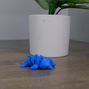 May include: A small, bright blue sculpture of a creature, possibly a sea animal, lies on a wooden surface. The sculpture is in front of a white cylindrical pot with a green plant. The sculpture's details are visible.