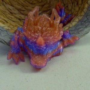 May include: A small, colorful dragon figurine in shades of red, orange, and blue. The dragon is lying down, with its head and body visible. The figurine has a textured surface and is set against a neutral background.