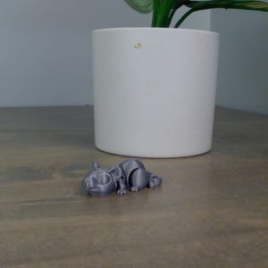 May include: A small, gray 3D-printed cat figurine lying on a wooden surface. The cat is in a relaxed pose, with its legs extended. A white cylindrical planter with a green plant is in the background.