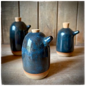 May include: Three ceramic oil bottles with a dark blue glaze and a natural cork stopper. The bottles have a spout and a light brown base. The background is a wooden surface.