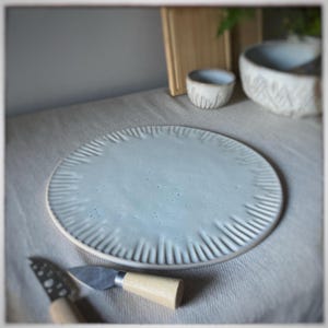May include: A pale blue ceramic plate with a textured edge, accompanied by two cheese knives with wooden handles. A small ceramic bowl and a larger patterned bowl are in the background, suggesting a dining setting.