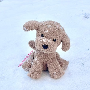 May include: A brown crocheted plush dog toy sitting in the snow. The dog has black eyes and a black nose.