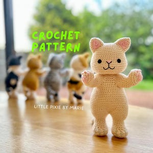 May include: Crochet pattern for a beige cat amigurumi toy. The cat is standing with its arms raised and has a cute face with black eyes and a pink nose. The text "CROCHET PATTERN" is written in green letters above the cat. The text "LITTLE PIXIE BY MARIE" is written below the cat.