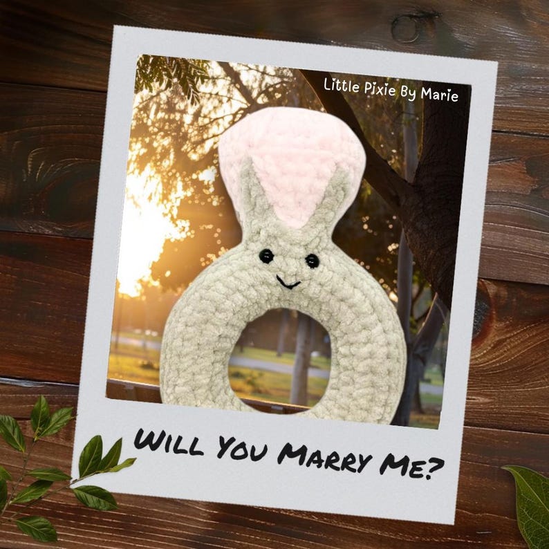 May include: A gray and pink plush ring with a smiling face and bunny ears. The ring is in a photo frame with the text "Will you marry me?" and "Little Pixie By Marie".