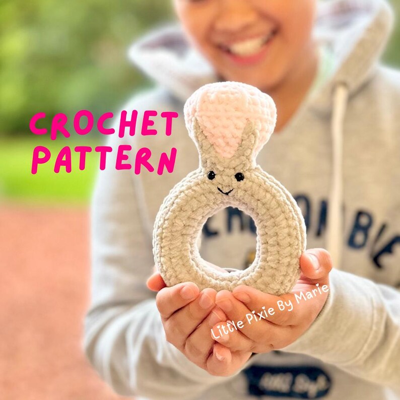 May include: A crocheted bunny ring toy with a pink nose and ears. The toy is held by a child's hands. The text "CROCHET PATTERN" is in pink letters on a white background. The text "Little Pixie By Marie" is written on the toy.