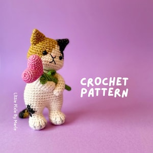 May include: Crochet pattern for a calico cat holding a pink heart-shaped flower. The cat is white with black and orange patches. The pattern is called "Little Pixie by Marie".