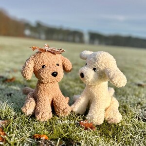 May include: Two plush toy dogs, one brown and one white, sitting on a bed of green grass with frost. The brown dog has a leaf on its head.