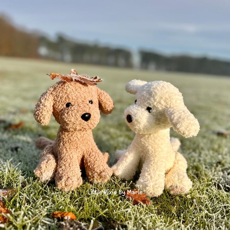 May include: Two plush toy dogs, one brown and one white, sitting on a bed of green grass with frost. The brown dog has a leaf on its head.