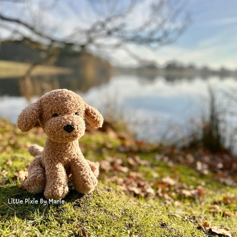 May include: A brown crocheted stuffed dog sitting on a bed of green moss with a blurry background of a lake and trees.