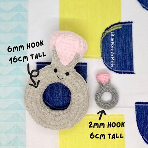 May include: Two crocheted ring toys. The larger ring is gray with a pink top and is 6 mm hook and 16 cm tall. The smaller ring is gray with a pink top and is 2 mm hook and 6 cm tall. The text "Little Pixie By Manie" is in the upper right corner of the image.
